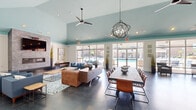 Located in Walkertown, NC, this thoughtfully designed single-floor community clubhouse offers a generous and versatile open-plan layout ideal for gathering and socializing. The space is immediately distinguished by its soaring vaulted ceilings painted in a striking aqua tone, which lend an airy, uplifting ambiance throughout. Polished concrete floors flow seamlessly across the entire space, providing a sleek, modern foundation that ties all areas together.

The main lounge area features a comfortable seating arrangement with plush blue armchairs, a leather ottoman, and a glass-topped coffee table, all anchored by a patterned area rug. A stacked stone fireplace with an integrated media console adds warmth and architectural character. Large floor-to-ceiling windows and glass doors flood the space with natural light and offer direct views of the outdoor pool area.

A long communal dining table with leather-and-metal chairs provides an inviting setting for events or casual gatherings. A glass-walled private office with a sliding door offers a quieter workspace within the open environment. Colorful abstract artwork adorns the white walls, adding creative energy throughout.

The fully equipped kitchenette features granite countertops, rich dark cabinetry, a coffee station, under-counter refrigerator, and a double sink — ideal for hosting events. A sleek orbital chandelier and modern ceiling fans complete the refined, contemporary aesthetic of this versatile community space.