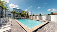 This contemporary residential community showcases a well-appointed outdoor amenity space designed for relaxed Florida living. The centerpiece is a generously sized swimming pool with crystal-clear blue water, framed by clean white perimeter walls and a tasteful paver deck that adds both visual appeal and practical functionality. Mature palm trees and lush tropical greenery surround the pool area, providing natural shade and a resort-like atmosphere beneath open, sun-filled skies.

Adjacent to the pool, a spacious patio area offers an inviting outdoor dining experience, complete with stylish bar-height wood tables and cushioned seating — ideal for entertaining or unwinding. The patio overlooks a well-maintained green lawn area and benefits from sweeping views of the surrounding landscape, including mature shade trees that contribute to a tranquil, park-like setting.

The building itself presents a clean, modern exterior with a light-toned facade, balconies, and large windows that reflect a contemporary architectural sensibility. The property is clearly designed with community and outdoor living in mind, seamlessly blending functional amenity spaces with attractive landscaping. The overall ambiance is bright, airy, and welcoming, characteristic of a thoughtfully designed Florida residential community.