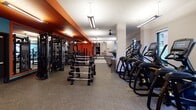 Located at 90 New Street in New Brunswick, NJ, this fitness center offers a comprehensive workout environment designed to accommodate a wide range of exercise needs. The facility is organized across a single floor, with distinct zones dedicated to cardio training, strength machines, and free weights.

The main workout area features a bold design aesthetic, with a striking burnt-orange accent ceiling running along the strength training zone, contrasting against clean white walls and structural columns. Durable speckled rubber flooring spans the space, providing both safety and practicality underfoot. Overhead, a combination of linear pendant lighting and ceiling fans ensures the environment remains well-lit and comfortable throughout.

The cardio section houses a row of modern Matrix treadmills and elliptical machines, positioned alongside windows that offer views of the surrounding New Brunswick cityscape, providing natural light and an open feel. A dedicated strength training area lines the perimeter with cable machines, selectorized equipment, and benches, while a centrally located dumbbell rack provides convenient access for free weight exercises.

A separate strength machine room features additional cable and selectorized machines alongside large windows, contributing to a bright and airy atmosphere. Overall, the facility presents a functional, modern, and energetic environment suited for a full-body fitness routine.