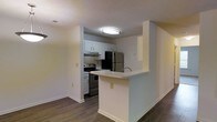 This single-floor residence offers a practical and well-designed layout with a seamless flow between living and kitchen spaces. The kitchen is a standout feature, outfitted with stainless steel appliances — including a refrigerator and range — white cabinetry, and light-toned countertops that provide ample prep space. A peninsula breakfast bar extends from the kitchen, creating an open pass-through that connects the cooking area to the living space, making it ideal for casual dining and entertaining. The kitchen also includes an under-cabinet range hood, adding to its functional design. Rich, dark hardwood-style flooring runs throughout the main living areas, offering a warm contrast to the bright white walls and cabinetry. The textured ceiling adds architectural character, while flush-mounted lighting ensures the space is well-lit throughout. Clean white baseboards and neutral wall tones give the interior a cohesive, move-in ready appearance. Overall, the space presents a clean, modern aesthetic with thoughtful design details that prioritize both comfort and functionality.