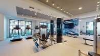 Located at 19400 US Highway 19 N in Clearwater, Florida, this single-level fitness center is designed to accommodate a wide range of workout routines in a polished, contemporary environment. The space is thoughtfully divided into distinct zones, each serving a specific fitness purpose while maintaining a cohesive aesthetic throughout.

The main workout area features a robust selection of strength and resistance machines, including a multi-functional cable system and weight benches, all arranged to maximize usability and flow. Track lighting runs along the ceiling, casting bright, even illumination across the light-toned carpeted floors. A striking large-scale abstract mural in shades of blue anchors one wall, complemented by a wall-mounted flat-screen television that adds both function and visual interest.

A dedicated cardio zone houses multiple treadmills positioned directly in front of expansive floor-to-ceiling windows framed in dark metal, offering views of palm trees and the surrounding landscape — an energizing backdrop for any workout. A separate studio room with warm wood flooring provides space for rowing and functional training, accented by colorful artwork and accessible via black-framed glass partition doors. Deep navy accent walls near the entrance lend a bold, modern contrast to the otherwise light and airy interior.