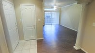 This single-story space offers a clean, functional layout centered around an open living area. The flooring transitions from light ceramic tile near the entryway to dark hardwood-style planks throughout the main living space, creating a clear distinction between zones. A paneled white door near the entry adds a classic architectural touch, complementing the neutral beige walls and white baseboards that run throughout.

The living area is bright and airy, benefiting from a large sliding glass door dressed with vertical blinds, which allows generous natural light to flood the interior while providing access to the outdoors. A ceiling fan is centrally positioned above the living space, offering both practical air circulation and a subtle decorative element. Crown molding details are visible along the upper walls, adding a refined finishing touch to the overall design.

The overall ambiance of the space is neutral and understated, with a palette of warm beiges and whites that lend themselves well to a variety of furnishing styles. The combination of durable flooring materials and simple architectural details makes this a practical and versatile living environment.