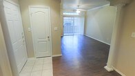 This single-story space offers a clean, functional layout centered around an open living area. The flooring transitions from light ceramic tile near the entryway to dark hardwood-style planks throughout the main living space, creating a clear distinction between zones. A paneled white door near the entry adds a classic architectural touch, complementing the neutral beige walls and white baseboards that run throughout.

The living area is bright and airy, benefiting from a large sliding glass door dressed with vertical blinds, which allows generous natural light to flood the interior while providing access to the outdoors. A ceiling fan is centrally positioned above the living space, offering both practical air circulation and a subtle decorative element. Crown molding details are visible along the upper walls, adding a refined finishing touch to the overall design.

The overall ambiance of the space is neutral and understated, with a palette of warm beiges and whites that lend themselves well to a variety of furnishing styles. The combination of durable flooring materials and simple architectural details makes this a practical and versatile living environment.