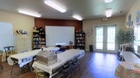 This single-floor community space is thoughtfully arranged to serve multiple purposes, from shared meals to leisure and learning. The main room features hardwood-style flooring that runs throughout, lending a warm and grounded feel to the interior. Flush-mounted fluorescent lighting panels illuminate the space evenly from above, ensuring a bright and functional environment.

Folding tables dressed with white tablecloths are set up in the center of the room, accompanied by folding chairs, and equipped with stainless steel chafing dishes — indicating the space is well-suited for communal dining and catered events. Along the walls, multiple tall bookshelves are stocked with a generous collection of books and are accented with decorative stuffed animals, adding a touch of whimsy and approachability.

The room benefits from natural light entering through a large window fitted with white horizontal blinds, as well as through a set of white-framed French doors that open to the exterior, providing both ventilation and a connection to the outdoors. An accent wall painted in a deep, warm red-brown tone adds character and visual depth to the space. A clearly marked exit sign and waste bin reflect the room's commitment to safety and cleanliness. Overall, this space presents as a versatile, community-oriented gathering area.