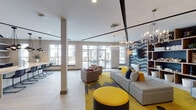 Located at 101 E Fourth St in Newport, Kentucky, this single-floor community lounge space delivers a striking contemporary aesthetic defined by bold design choices and thoughtfully curated amenities. The open-plan layout is anchored by a vibrant seating area adorned with gray modular sofas, accent chairs, and eye-catching yellow ottomans atop a vivid yellow-and-gray patterned area rug. A standout feature wall clad in a graphic black-and-white geometric wallcovering frames a wall-mounted flat-screen TV, flanked by a substantial rustic wood open shelving unit. Elegant multi-arm chandeliers hang above the lounge area, complementing the recessed lighting throughout the space. An expansive island-style kitchen bar with sleek dark cabinetry, a stone countertop, and bar seating serves as a social and functional hub, illuminated by modern pendant cluster lighting. Large floor-to-ceiling windows flood the interior with abundant natural light and provide views of an outdoor terrace fitted with additional seating. Toward the rear of the space, a game area featuring a billiards table adds a recreational element. Light-toned tile flooring and clean white ceilings with contrasting dark accent trim lines complete the polished, urban atmosphere of this well-appointed gathering space.