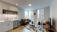 This well-appointed one-bedroom unit is thoughtfully designed with a contemporary aesthetic and a practical, open-concept layout. Light wood-tone flooring flows consistently throughout the entire space, contributing to a clean, cohesive look.

The living area serves as the heart of the unit, anchored by floor-to-ceiling corner windows that flood the room with natural light and frame expansive urban skyline views. The generous window placement creates an airy, open atmosphere while connecting the interior to the city beyond.

The kitchen is efficiently arranged in an L-shaped configuration, featuring flat-front cabinetry in a soft gray-white finish, white quartz countertops, a subway tile backsplash, and a full suite of stainless steel appliances including a refrigerator, range, microwave, and dishwasher. Open shelving adds a modern, functional touch.

The bedroom is a calm, restful space with soft neutral walls, a large window with urban views, and a reach-in closet with sliding doors providing ample storage.

The bathroom is finished in warm taupe tile surrounding a full soaking tub and shower combination, complemented by a white vanity, wood-look flooring, and glass shelving, creating a clean and modern atmosphere throughout.