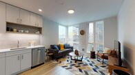 This well-appointed one-bedroom unit is thoughtfully designed with a contemporary aesthetic and a practical, open-concept layout. Light wood-tone flooring flows consistently throughout the entire space, contributing to a clean, cohesive look.

The living area serves as the heart of the unit, anchored by floor-to-ceiling corner windows that flood the room with natural light and frame expansive urban skyline views. The generous window placement creates an airy, open atmosphere while connecting the interior to the city beyond.

The kitchen is efficiently arranged in an L-shaped configuration, featuring flat-front cabinetry in a soft gray-white finish, white quartz countertops, a subway tile backsplash, and a full suite of stainless steel appliances including a refrigerator, range, microwave, and dishwasher. Open shelving adds a modern, functional touch.

The bedroom is a calm, restful space with soft neutral walls, a large window with urban views, and a reach-in closet with sliding doors providing ample storage.

The bathroom is finished in warm taupe tile surrounding a full soaking tub and shower combination, complemented by a white vanity, wood-look flooring, and glass shelving, creating a clean and modern atmosphere throughout.
