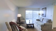This single-floor residential space offers a warm and comfortable living environment designed for everyday living. The living room serves as the heart of the home, featuring plush wall-to-wall carpeting that adds a soft, inviting feel underfoot. A large sliding glass door, dressed with vertical blinds, acts as a focal point for the room, flooding the space with abundant natural light and providing access to an outdoor area. The room accommodates a well-arranged seating arrangement, including a sofa and accent chair, centered around a dark wood coffee table that adds a touch of classic elegance. Decorative table lamps on either side of the seating area contribute to a cozy, layered lighting scheme. The textured ceiling adds a subtle architectural detail throughout. Overall, the space presents a functional and comfortable atmosphere, balancing natural light with traditional furnishings to create a lived-in, welcoming environment suited for relaxing and entertaining.