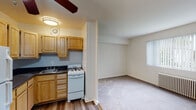 This single-floor unit offers a practical and straightforward layout designed to maximize functionality. The kitchen is equipped with light wood cabinetry providing ample storage, dark countertops, a double sink, a gas range, and a full-size refrigerator. Wood-style flooring runs throughout the kitchen area, while a ceiling-mounted light fixture and ventilation fan ensure proper illumination and airflow.

Adjacent to the kitchen, the living area features neutral carpet and a ceiling fan, creating a comfortable, open feel. A window with vertical blinds allows natural light to filter in. A separate carpeted room, likely a bedroom, includes a radiator heating unit, a window-mounted air conditioning unit, and a window overlooking the exterior.

A generously sized walk-in closet offers extensive built-in shelving and hanging space along multiple walls, providing notable storage capacity.

The bathroom features a classic retro aesthetic with cream and black tile work lining the walls, a soaking tub with a shower, a white vanity with mirror, and tiled flooring. Windows in the bathroom allow for natural light. Overall, the unit maintains a clean, neutral palette throughout.