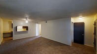 This single-floor residential unit offers a straightforward and functional layout designed for comfortable everyday living. The main living area is generously sized, featuring wall-to-wall carpet flooring in a warm neutral tone that adds a cozy feel underfoot. A ceiling fan is centrally mounted overhead, providing both lighting and air circulation throughout the space. The textured popcorn ceiling adds a classic residential character to the room.

The open layout creates a seamless visual connection between the living area and the kitchen, which is visible in the background and features dark wood cabinetry paired with a lighter countertop surface, offering a practical workspace. A doorway leading to additional areas of the unit — likely bedrooms or a hallway — is accessible from the main living space, hinting at a well-connected floor plan.

The walls are finished in a clean, neutral white, providing a blank canvas that suits a variety of décor styles. A visible HVAC vent in the ceiling suggests a central climate control system. The transition to wood-style flooring is visible near the kitchen and hallway areas, adding subtle contrast to the carpeted main living zone. Overall, the space presents a clean, move-in-ready residential environment with practical features throughout.