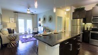 This single-floor residence offers a thoughtfully designed open-concept layout that seamlessly connects the kitchen and living areas, creating a spacious and inviting atmosphere. The kitchen is a standout feature, anchored by a large island with a striking dark granite countertop, dark espresso cabinetry with sleek hardware, and elegant pendant lighting hanging above. Stainless steel appliances add a modern, polished touch to the space.

Rich hardwood flooring flows throughout, adding warmth and continuity from the kitchen into the living area. The living space is generously sized, bathed in natural light that streams through French doors leading to the outside. A ceiling fan overhead ensures comfort year-round, while soft, neutral wall tones and tasteful décor accents — including sunburst wall art — contribute to the refined aesthetic.

The overall ambiance is both contemporary and comfortable, with a well-balanced blend of dark and light tones that create visual depth. The open sightlines between rooms enhance the sense of spaciousness, making this residence ideal for both everyday living and entertaining.