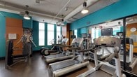 This fitness center offers a thoughtfully designed workout environment that balances functionality with modern aesthetics. The space is defined by striking teal accent walls paired with warm wood-toned paneling, creating a vibrant yet focused atmosphere for exercise. The industrial-style ceiling features exposed ductwork and conduit alongside pendant and track lighting, lending the room an urban, contemporary character.

The cardio area is well-stocked with a row of treadmills positioned toward the front, complemented by elliptical trainers and additional cardio equipment arranged throughout the space. Each machine is equipped with its own personal monitor, allowing users to stay entertained or track their workouts conveniently. Large windows along one wall admit natural light, brightening the interior while providing a visual connection to the outside.

A mirrored wall extends along one side of the room, enhancing the sense of space and allowing users to monitor their form during exercise. The flooring consists of durable rubber matting, practical for a high-traffic fitness environment. Overall, the space is clean, organized, and well-suited to support a comprehensive cardio-focused workout routine.