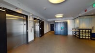 Located at 928 N Western Ave in the East Hollywood neighborhood of Los Angeles, this ground-floor lobby and mail area serves as the central point of entry and resident services for the building. The space is designed with a clean, modern aesthetic, featuring smooth gray walls, recessed lighting, and durable stone-tiled flooring that transitions to carpet in the main corridor.

The elevator lobby is a key feature, housing two stainless steel elevator doors with dark-framed surrounds, providing convenient vertical access throughout the building. Call buttons and informational signage are neatly mounted between the elevators, reflecting a well-organized and functional layout.

Adjacent to the elevators is a dedicated package management area equipped with an Amazon Hub smart locker system, offering residents a secure and convenient way to receive deliveries around the clock. The locker unit features a digital touchscreen interface and is prominently positioned for easy access.

A separate mail vestibule near the building's front entrance houses an extensive array of individual resident mailboxes lining the walls. This area is accessed through a decorative glass and metal security door, which features a geometric square-pattern design that adds an architectural detail to the entry. Recessed lighting and an emergency exit sign provide both ambiance and safety compliance. Overall, the lobby presents a practical yet polished environment suited to a multi-unit residential building.