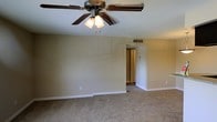 This single-story space presents a clean, straightforward layout designed for comfortable everyday living. The main living area is finished with wall-to-wall plush carpeting in a neutral beige tone, complementing the warm, light-colored walls that create a calm and inviting atmosphere throughout.

A ceiling fan with an integrated multi-bulb light fixture serves as a central overhead feature, providing both air circulation and ambient lighting to the room. The smooth white ceiling and coordinating white baseboards add a crisp, well-maintained appearance to the space.

A centrally positioned open doorway connects the main living area to the adjacent hallway and additional rooms beyond, allowing for natural flow and accessibility between spaces. A wall-mounted thermostat is visible near the passageway, and a ceiling-mounted HVAC vent ensures consistent climate control throughout. Electrical outlets are conveniently placed along the walls.

Overall, the space offers a simple, functional design with neutral finishes that provide a versatile backdrop, making it easy to personalize with furnishings and décor to suit a variety of lifestyles.
