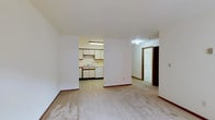 This single-floor residential space offers a practical and straightforward layout designed for comfortable everyday living. The main living area is finished with wall-to-wall carpeting in a neutral tone, creating a warm and understated foundation that pairs well with the clean white walls and wood-accented baseboards throughout.

Seamlessly connected to the living area is a functional kitchen, separated by a partial wall that maintains an open feel between the two spaces. The kitchen features white cabinetry with ample storage, a double-basin sink, a dishwasher, and tile flooring that contrasts neatly with the carpeted areas. Overhead fluorescent lighting ensures the kitchen is well-lit and practical for daily use.

A hallway extends from the living area toward additional rooms, hinting at a bathroom and further private spaces beyond. The hallway is carpeted and finished consistently with the rest of the unit, contributing to a cohesive interior aesthetic.

Overall, the space presents a clean, move-in-ready environment with a simple and functional design. The neutral color palette throughout provides a versatile backdrop, making it easy to personalize with furnishings and décor. The layout is efficient, with each area clearly defined while maintaining an open and connected flow.