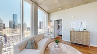 This contemporary high-rise unit is defined by its striking industrial-modern architecture, showcasing exposed concrete ceilings throughout and wide-plank hardwood flooring that flows seamlessly across all living spaces. Floor-to-ceiling windows are a hallmark of the unit, flooding every room with abundant natural light and offering panoramic views of the surrounding urban skyline and cityscape below.

The open-concept kitchen and living area form the heart of the space. The kitchen is outfitted with stainless steel appliances, two-tone cabinetry in white and warm wood tones, a subway tile backsplash, and track lighting mounted to the concrete ceiling. It connects directly to the living area, which benefits from dramatic corner window views and access to a small dining nook.

A dedicated den or secondary sitting room provides versatile living space, complete with a built-in arched wooden bookshelf, a floor-to-ceiling window framing expansive city views, and a cozy seating arrangement. An entry hallway with clean white walls leads naturally into the main living areas.

The bedroom is generously proportioned, featuring floor-to-ceiling windows on two walls that capture sweeping skyline panoramas, an exposed concrete ceiling, and a walk-in closet with built-in shelving. The full bathroom is refined and spa-like, with a double-sink vanity topped in white quartz, warm wood cabinetry with brushed gold hardware, an LED-lit mirror, and a full glass-enclosed walk-in shower clad in large-format stone tile.