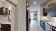 This single-floor residence showcases a contemporary design with light hardwood-style flooring and recessed lighting carried consistently throughout. The open-concept layout connects the living area directly to the kitchen, creating a seamless flow between spaces. The galley-style kitchen is well-appointed with dark cabinetry, granite countertops, a white subway tile backsplash, and a full suite of stainless steel appliances including a refrigerator, range, built-in microwave, and dishwasher. A contrasting section of blue cabinetry with granite counters adds a distinctive design accent along one wall.

The living area is bright and open, with recessed lighting overhead and direct access to a private balcony through a glass door, allowing natural light to filter in. Built-in shelving provides functional storage within the room, complemented by a ceiling fan for added comfort.

A dedicated mudroom-style nook near the balcony entry features built-in cubbies and shelving, offering practical organization space. The bathroom maintains the cohesive design aesthetic with dark wood cabinetry, granite countertops, a large vanity mirror, and a tiled walk-in shower. Clean white walls and consistent flooring throughout contribute to a bright, modern ambiance.