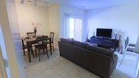This single-floor space features an open-concept layout that connects the living and dining areas, creating a cohesive and functional environment. The living area is anchored by a large sectional sofa positioned to face a wall-mounted flat-screen television set atop a low media console. Plush carpeted flooring runs throughout, adding warmth and comfort underfoot. A sliding glass door fills the space with natural light and provides access to an outdoor balcony area, serving as a bright focal point in the room. The dining area is situated adjacent to the living space and accommodates a dark wood dining table paired with matching bar-height chairs, making it well-suited for casual meals or gatherings. A decorative floral arrangement adds a touch of color to the dining corner. Track lighting installed along the ceiling in the dining area ensures practical and adjustable illumination. The walls are finished in a neutral, light tone that enhances the brightness of the space. Overall, the layout is efficient and well-organized, offering a comfortable living environment with clearly defined functional zones within an open, connected floor plan.