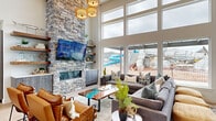 This impressive single-floor communal space is designed to make a bold architectural statement from the moment you enter. The entry foyer features light hardwood flooring, high clerestory windows, and frosted glass panels that lend a polished, welcoming feel. At the heart of the space, a dramatic floor-to-ceiling stacked stone fireplace commands attention, flanked by open wood shelving and a wall-mounted television — a centerpiece that anchors the main lounge area. Soaring two-story windows flood the space with natural light and offer unobstructed views of an outdoor water park and recreation area beyond, creating a vibrant indoor-outdoor connection. The lounge is furnished with generous sectional seating, leather accent chairs, and a striking live-edge resin coffee table, all set atop light hardwood floors. Woven pendant and drum chandelier lighting add warmth and texture overhead. A dining area with round dark-wood tables and leather chairs provides additional seating, while a fully equipped kitchenette — outfitted with textured grey cabinetry, stainless steel appliances, and a modern pull-down faucet — adds everyday functionality. The overall ambiance is contemporary and resort-inspired, balancing comfort with distinctive design.
