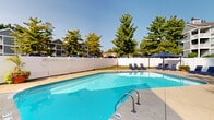 Located at 5680 Hibernia Dr in Columbus, Ohio, this outdoor pool area serves as a central community amenity designed for relaxation and recreation. The centerpiece is a generously sized swimming pool with clear, blue water, accessible via stainless steel entry rails and surrounded by a wide concrete sundeck that provides ample space for lounging and socializing.

The deck is furnished with multiple navy blue chaise lounge chairs, thoughtfully arranged around the pool's perimeter to maximize sun exposure and comfort. A freestanding umbrella offers shaded seating for those seeking relief from the sun. The entire area is enclosed by a tall white vinyl privacy fence, creating a secure and peaceful environment.

Adjacent to the pool is a single-story poolside structure with a pitched roof and lattice detailing, which appears to house a utility or storage area. Safety equipment, including a life ring, is visibly posted near the structure, reflecting a commitment to pool safety standards.

Mature evergreen and deciduous trees line the perimeter, providing a lush, green backdrop and natural shade. The surrounding landscape enhances the sense of a private retreat, with well-maintained grassy areas adding to the overall appeal. The space is open to the sky, allowing for abundant natural light throughout the day, making it an inviting outdoor destination across the warmer months.