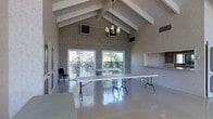This single-floor space is designed to accommodate gatherings and events, offering an open and versatile layout. The interior is immediately defined by its dramatic vaulted ceiling, accented with exposed white wooden beams and white-painted shiplap paneling that runs along the ceiling's pitch — creating a sense of considerable vertical volume and architectural character.

A full-height brick accent wall serves as a striking focal point, anchoring one end of the main hall and adding a rustic, textural contrast to the otherwise clean and neutral interior. A classic chandelier hangs at the apex of the vaulted ceiling, providing ambient overhead lighting that complements the natural light streaming in through large sliding glass doors and panel windows along the exterior wall.

These glass doors open to an outdoor area visible beyond lattice fencing, allowing natural light to flood the interior and offering a seamless connection to the outdoors. The polished concrete floors throughout reflect light effectively, enhancing the sense of spaciousness and making the area easy to maintain.

A built-in pass-through counter or service window is visible along one wall, suggesting utility for catering or refreshment service during events. The space is currently furnished with long folding tables, underscoring its adaptability for a wide range of uses including meetings, receptions, or community events.