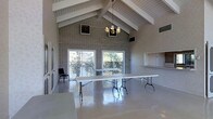 This single-floor space is designed to accommodate gatherings and events, offering an open and versatile layout. The interior is immediately defined by its dramatic vaulted ceiling, accented with exposed white wooden beams and white-painted shiplap paneling that runs along the ceiling's pitch — creating a sense of considerable vertical volume and architectural character.

A full-height brick accent wall serves as a striking focal point, anchoring one end of the main hall and adding a rustic, textural contrast to the otherwise clean and neutral interior. A classic chandelier hangs at the apex of the vaulted ceiling, providing ambient overhead lighting that complements the natural light streaming in through large sliding glass doors and panel windows along the exterior wall.

These glass doors open to an outdoor area visible beyond lattice fencing, allowing natural light to flood the interior and offering a seamless connection to the outdoors. The polished concrete floors throughout reflect light effectively, enhancing the sense of spaciousness and making the area easy to maintain.

A built-in pass-through counter or service window is visible along one wall, suggesting utility for catering or refreshment service during events. The space is currently furnished with long folding tables, underscoring its adaptability for a wide range of uses including meetings, receptions, or community events.