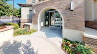 This single-story commercial building presents a polished and welcoming exterior characterized by a prominent arched entryway framed in warm brick and smooth stucco. The entrance is anchored by glass double doors with accessible entry provisions, flanked by wall-mounted lighting fixtures that add a refined touch to the façade. The exterior walls feature a combination of textured brick and stucco finishes that lend the building a professional yet approachable aesthetic.

The surrounding grounds are thoughtfully landscaped, incorporating a variety of ornamental plants, flowering shrubs, and mature trees that provide seasonal color and natural shade. Curved concrete planters and low seating walls accent the entry approach, guiding visitors along a broad, smooth-paved walkway that sweeps gracefully toward the entrance. The outdoor areas benefit from abundant natural sunlight, creating a bright and inviting atmosphere throughout the day.

The building is situated within a well-developed mixed-use environment, with paved parking areas, decorative paver driveways, and neighboring multi-story residential structures visible in the surrounding landscape. Mature trees line the perimeter, offering greenery and softening the built environment. The overall setting conveys a sense of order, accessibility, and community integration, making this an appealing and functional space within its broader neighborhood context.