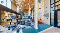 This impressive two-floor fitness facility in Morrisville, NC is designed to deliver a comprehensive workout experience across a generous total footprint. The main floor immediately captivates with soaring double-height ceilings adorned with decorative globe pendant lighting and a bold geometric-patterned carpet that flows throughout. Floor-to-ceiling windows flood the space with natural light and offer views of the surrounding landscape, while exposed stone accent walls and a large-scale athletic mural contribute to the dynamic, energetic ambiance. A standout rock climbing wall anchors one corner of the main floor, complemented by an array of cardio machines including treadmills and stair climbers. A sculptural spiral staircase with wood-and-steel detailing connects the two levels. The upper floor houses additional exercise areas, including a cycling room with smart bikes, a stretching and functional training zone with resistance bands and accessory equipment, and access to an outdoor balcony. A dedicated weight training room features hardwood floors, rack systems, benches, dumbbells, cable machines, full-length mirrors, and built-in display cabinetry. Sleek linear lighting and a clean, modern aesthetic define this space throughout.