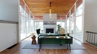 This single-story space showcases a bold mid-century modern aesthetic defined by architectural precision and an seamless connection to the outdoors. The living area is the centerpiece, featuring dramatically vaulted ceilings clad in warm natural wood planking, which add richness and texture overhead. Floor-to-ceiling glass walls envelop the space on multiple sides, flooding the interior with abundant natural light and offering sweeping views of the surrounding mature trees and outdoor areas, creating an immersive indoor-outdoor atmosphere.

A full-height stacked stone fireplace serves as the room's focal anchor, its light-toned facade extending to the ceiling and topped with a curated gallery of framed artwork along the mantel. Warm hardwood flooring runs throughout, complementing the wood ceiling and grounding the space with continuity. Furnishings follow the mid-century theme, including tufted green velvet seating, hairpin-leg accent tables, and a sculptural pendant light cluster above.

The kitchen features white shaker-style cabinetry, speckled granite countertops, and stainless steel appliances, offering a clean and functional layout. A large sliding glass door connects the living area directly to an adjacent sunlit space, enhancing the home's open, airy character throughout.