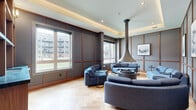Located at 2274 Princess St in Kingston, Ontario, this single-floor lounge space offers a refined and thoughtfully designed interior that blends modern elegance with warm, inviting details. The main living area is anchored by a dramatic suspended cone-shaped fireplace that descends from a coffered tray ceiling, serving as a striking architectural focal point. Surrounding it, curved blue upholstered sofas and armchairs are arranged to encourage conversation, all resting on beautifully patterned herringbone hardwood flooring that extends throughout the space. The walls are finished with a grid of wood-framed panels in warm tones against a cool grey backdrop, lending a structured yet sophisticated aesthetic. Large windows flood the room with natural light while offering views of the surrounding outdoor courtyard. An adjacent area, enclosed by sleek black-framed glass partition doors, houses a dedicated wine cellar complete with floor-to-ceiling wooden lattice wine racks and a round dining table, ideal for intimate gatherings. Recessed lighting and subtle wall sconces complement the tray ceiling's indirect lighting, creating a warm, layered ambiance. The entry corridor connects seamlessly to the main lounge, maintaining a cohesive design language throughout. This space offers an elevated communal environment with exceptional attention to architectural and interior detail.