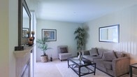 This single-floor space presents a well-appointed living area designed with a clean, contemporary aesthetic. The living room is finished with soft, light green walls that create a calm and welcoming atmosphere, complemented by plush wall-to-wall carpeting underfoot. Natural light filters into the space, enhancing the airy and open feel of the room. The layout comfortably accommodates a full seating arrangement, including a generously sized sofa with accent pillows and a coordinating patterned accent chair with an ottoman, both centered around a sleek glass-top coffee table with a dark metal frame. Framed artwork adorns the walls, adding visual interest without overwhelming the understated palette. Tall indoor plants placed in the corners bring a natural, organic element to the space, contributing to a fresh and lively ambiance. The overall design balances simplicity and comfort, making the living area feel both functional and inviting. The neutral color scheme and minimal décor allow for easy personalization and flexibility in furnishing styles.