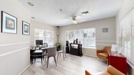 This single-floor commercial space is designed to offer a professional yet inviting atmosphere for both staff and visitors. The main open area serves as a combined reception and workspace, featuring light hardwood-style flooring, a textured ceiling with a ceiling fan, and white horizontal blinds across multiple large windows that allow abundant natural light to fill the room. A decorative chair rail runs along the walls, lending a polished, structured look throughout.

The primary workstation area is equipped with a sleek black desk, ergonomic seating, and a computer setup, positioned to take advantage of the natural light from the surrounding windows. A private office, accessible through a glass-paned French door, offers a dedicated enclosed workspace complete with a desk, additional seating, and framed artwork on the walls.

A welcoming reception corner features a vibrant red display cabinet stocked with snacks and goods, alongside a cognac leather accent chair — creating a warm, comfortable waiting area. A beverage station with a coffee maker adds to the hospitality of the space.

Adding a fun and memorable touch, a classic red popcorn cart is stationed in a nook adorned with framed artwork, contributing to the space's approachable and personable character. Throughout, the neutral warm-beige walls, consistent flooring, and thoughtful furnishings create a cohesive, professional, and welcoming environment.