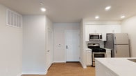 This single-floor space showcases a clean, contemporary design with a seamless open-concept layout that connects the entryway and kitchen into one cohesive living area. The entryway is bright and welcoming, featuring light hardwood-style flooring that extends throughout the space, crisp white walls, and recessed lighting that provides even, warm illumination across the ceiling.

The kitchen is positioned adjacent to the entry and is well-equipped with stainless steel appliances, including a gas range and an over-the-range microwave. White shaker-style cabinetry lines the upper and lower walls, offering ample storage, while light-colored countertops contribute to the space's bright, airy feel. A louvered door near the entryway likely conceals a utility closet or storage area, adding practical functionality to the layout.

The recessed lighting throughout ensures consistent brightness, complementing the neutral color palette of white and light gray tones that define the overall aesthetic. The space presents a modern, well-maintained environment with thoughtful finishes and a straightforward, functional design suited for everyday living.