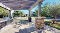 Located at 18545 University Blvd in Sugar Land, TX, this outdoor pergola pavilion offers a well-appointed communal gathering space designed for comfort and functionality. The structure is anchored by substantial stacked-stone columns that lend a refined, rustic character to the space, supporting a slatted wood overhead pergola that provides partial shade while allowing natural light and airflow throughout.

A standout feature of the pavilion is the built-in outdoor kitchen station, complete with a stainless steel gas grill set into a matching stacked-stone surround — ideal for gatherings and cookouts. Ceiling fans mounted along the pergola ensure comfort during warm Texas days, while decorative wall-mounted lanterns add a touch of ambiance. A concrete patio floor provides a clean, durable surface underfoot, and a waste receptacle is conveniently positioned within the space.

The pavilion opens on multiple sides to expansive, manicured green lawns dotted with mature trees, offering pleasant views of the surrounding landscaped grounds and adjacent multi-story residential buildings. Concrete walkways connect the pavilion to the broader property, making it easily accessible. Colorful potted plants add decorative accents throughout. Overall, this outdoor space delivers a functional yet inviting environment for outdoor dining, socializing, and relaxation.