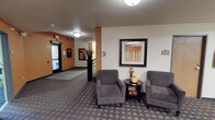 This single-floor residential unit is part of a larger multi-unit building, offering a functional and comfortable layout designed for everyday living. The building's shared lobby sets a welcoming tone, featuring patterned carpeting, upholstered seating, framed wall art, and warm accent lighting — creating an inviting entry point for residents and guests alike.

The unit itself is finished with neutral, light-colored walls and consistent wall-to-wall carpeting throughout, providing a clean and understated aesthetic. The main living area is generously sized and open, with white walls that allow for flexible furnishing arrangements. A large window fitted with vertical blinds fills the room with natural light and offers a pleasant view of surrounding greenery and trees, contributing to a calm and airy atmosphere.

The bedroom area provides ample storage through a dedicated walk-in closet with built-in shelving, along with additional closet access. The layout connects conveniently to a kitchen area, visible through an open doorway and featuring appliances including a refrigerator. Baseboard heating is present throughout, ensuring consistent warmth. Smoke detection is also installed for safety. The unit's straightforward, practical design makes it well-suited for comfortable, low-maintenance living.