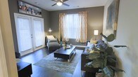 This single-story space showcases a thoughtfully designed living room that balances comfort and style. The room is anchored by rich, dark hardwood flooring that contrasts elegantly with the warm gray walls, creating a sophisticated yet inviting atmosphere. A ceiling fan positioned centrally overhead provides both function and a subtle architectural focal point.

Natural light is a defining feature, with multiple windows dressed in white horizontal blinds and accented by eye-catching orange and white patterned curtains that introduce a lively pop of color. The window placement allows light to filter generously throughout the room.

The living area is furnished with a gray sectional sofa adorned with yellow and teal accent pillows, complemented by a matching chaise lounge chair. A dark rectangular coffee table sits atop a neutral area rug, grounding the seating arrangement. Floor lamps positioned throughout provide warm ambient lighting for evening use.

Lush indoor plants add a natural, organic element to the décor, while decorative artwork displayed along the upper wall adds a personal, curated touch. The overall ambiance is modern, cohesive, and welcoming.