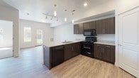 This single-story residence showcases a modern, open-concept design with a consistent neutral color palette and clean architectural lines throughout. The kitchen serves as the centerpiece of the living area, featuring dark espresso-toned cabinetry with sleek bar-pull hardware and light granite countertops. A peninsula with an integrated sink extends into the living space, creating a natural division while maintaining an open flow. Black appliances, including a range, microwave, and refrigerator, complement the cabinetry. Pendant lighting hangs above the peninsula, while recessed lighting and a track light fixture provide additional illumination. Light-toned luxury vinyl plank flooring runs throughout the main living areas, offering both durability and visual continuity. A door provides direct access to the outdoors, allowing natural light to filter into the space. The bedrooms are finished with wall-to-wall carpet, providing a softer underfoot feel, and feature windows that bring in natural light. The bathroom includes a full tub-shower combination, a granite-topped vanity with dark cabinetry matching the kitchen, a large mirror, and LVP flooring consistent with the rest of the unit. The overall aesthetic is clean, contemporary, and cohesive.
