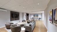 This single-floor amenity space is thoughtfully designed to offer a seamless blend of relaxation and recreation. The open-concept layout is anchored by a generously sized lounge area furnished with plush grey sofas, accent chairs, and a round coffee table, all arranged atop a patterned area rug over warm hardwood flooring. A large wall-mounted flat-screen television is set against a striking dark wood accent wall, creating a bold focal point that adds warmth and contrast to the otherwise neutral, light grey palette throughout.

Recessed lighting is embedded in a grid-panel ceiling, casting a clean, even illumination across the entire space. The layout flows naturally into a game room area at the rear, featuring a full-size billiards table and additional high-top seating — ideal for social gatherings. A vibrant cityscape artwork adorns the back wall, injecting a pop of color and visual interest to the room.

The overall ambiance is polished and contemporary, with a refined aesthetic that balances comfort with style. Clean lines, neutral tones, and quality furnishings give the space a professional yet inviting atmosphere, making it well-suited for both casual socializing and organized group activities.