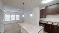 This single-floor residence offers a thoughtfully designed open-concept layout that seamlessly connects the kitchen and living areas. The kitchen is a standout feature, showcasing rich dark-stained cabinetry that provides ample storage, complemented by granite countertops with subtle natural veining. A spacious kitchen island serves as both a functional workspace and a casual gathering spot, anchored by two modern pendant lights that add warm, focused illumination. Stainless steel appliances complete the kitchen's contemporary aesthetic. The open layout flows naturally into the living area, where a ceiling fan with light fixture keeps the space comfortable and well-lit. Large windows dressed with horizontal blinds allow natural light to filter in, brightening the neutral-toned walls and light carpeted flooring. The overall ambiance is clean, modern, and inviting, with a cohesive color palette of whites, grays, and deep browns that lends the space a timeless, polished character. The single-floor plan ensures easy accessibility and a smooth, connected flow throughout the home.