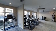 Located in Carrollton, Texas, this fitness center offers a thoughtfully designed, single-floor workout space that balances functionality with a clean, modern aesthetic. The interior is finished with neutral taupe walls and low-pile carpeting, creating a calm and focused environment for exercise.

The cardio area is a standout feature, lined with a row of modern elliptical trainers and additional cardio machines positioned to take full advantage of the large windows along the exterior walls. These windows flood the space with natural light while offering views of the surrounding exterior, enhancing the overall workout experience.

Structural columns divide the space subtly without disrupting the open flow of the layout. Recessed ceiling lighting supplements the natural light, ensuring the space remains well-lit throughout the day. Ceiling fans are installed overhead, promoting air circulation and comfort during intense workouts.

Additional strength and resistance equipment is visible toward the rear of the space, indicating a well-rounded facility capable of supporting diverse fitness routines. The overall ambiance is bright, airy, and organized, making it a practical and welcoming environment for daily fitness use.