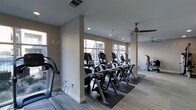 Located in Carrollton, Texas, this fitness center offers a thoughtfully designed, single-floor workout space that balances functionality with a clean, modern aesthetic. The interior is finished with neutral taupe walls and low-pile carpeting, creating a calm and focused environment for exercise.

The cardio area is a standout feature, lined with a row of modern elliptical trainers and additional cardio machines positioned to take full advantage of the large windows along the exterior walls. These windows flood the space with natural light while offering views of the surrounding exterior, enhancing the overall workout experience.

Structural columns divide the space subtly without disrupting the open flow of the layout. Recessed ceiling lighting supplements the natural light, ensuring the space remains well-lit throughout the day. Ceiling fans are installed overhead, promoting air circulation and comfort during intense workouts.

Additional strength and resistance equipment is visible toward the rear of the space, indicating a well-rounded facility capable of supporting diverse fitness routines. The overall ambiance is bright, airy, and organized, making it a practical and welcoming environment for daily fitness use.