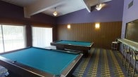 This single-floor billiards room is designed as a dedicated recreational space, offering ample room to accommodate two full-sized pool tables with turquoise felt surfaces. The layout provides comfortable spacing between the tables, allowing players to move freely and take shots without obstruction. The walls feature classic vertical wood paneling in a warm brown tone, adding a traditional and inviting character to the space. The upper portion of the walls is painted in a deep purple hue, creating a bold contrast against the paneling below and contributing to the room's distinct atmosphere. Overhead, pendant light fixtures hang from a coffered ceiling, casting a soft ambient glow, while wall-mounted sconces positioned along the wood-paneled walls provide additional warm, focused lighting. A large window with vertical blinds on one wall allows natural light to filter into the space during daytime hours. The floor is covered in a patterned carpet featuring a geometric design in muted tones of blue, green, and gold, adding visual interest underfoot. Overall, the room presents a well-appointed, purposeful environment suited for leisure and social activity.
