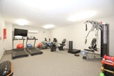 This fitness facility is designed to accommodate a well-rounded exercise experience across its single floor. The space is divided into distinct functional zones, each outfitted with purpose-driven equipment and finished with durable speckled flooring suited to high-traffic use. Neutral-toned walls and flush panel lighting throughout create a clean, focused environment conducive to physical training.

The cardio area features two treadmills by True and Spirit, positioned facing a wall-mounted flat-screen television, with a wall-mounted air conditioning unit ensuring climate comfort during use. An assortment of stability and medicine balls in various sizes is neatly arranged nearby, alongside stationary exercise bikes for low-impact cardiovascular training.

A separate strength-training section houses a multi-function cable and weight machine, an adjustable exercise bench, a BOSU balance trainer, and exercise mats for floor-based workouts. A dumbbell rack stocked with a range of weighted options provides versatility for resistance training, while medicine balls are also available for functional fitness routines.

A wall-mounted wooden balance rail is present, suggesting potential use for rehabilitation or balance exercises. An adjacent room accessible through a standard door appears to serve as a utility or storage space, adding practicality to the facility's overall layout.