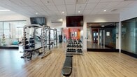 Located at 260 E Main St in downtown Rochester, NY, this modern fitness center offers a comprehensive workout facility spread across a single floor. The main gym area features warm wood-look flooring, a suspended drop ceiling with recessed lighting, and a vibrant red accent wall that adds energy to the space. Multiple flat-screen TVs are mounted throughout, and a wall-mounted air conditioning unit ensures a comfortable environment during workouts.

The strength training zone is well-appointed with a power rack, adjustable benches, a full dumbbell rack ranging across various weights, and a selection of cable and resistance machines. A mirrored wall expands the sense of space and allows for form checks during lifts.

The cardio section benefits from abundant natural light streaming through large windows that frame views of the surrounding downtown Rochester skyline. Equipment includes treadmills, stair climbers, and elliptical machines, all positioned to take advantage of the city views.

A dedicated spin studio provides a separate space for cycling classes, equipped with multiple stationary bikes, stability balls, and a wall-mounted TV. Large windows in this room also let in generous natural light, creating an bright and motivating atmosphere.

Together, these areas form a well-rounded, professionally outfitted fitness facility suited for a full range of exercise routines.