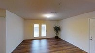 This single-story space presents a straightforward and functional interior anchored by rich, dark hardwood flooring that extends throughout the main living area. The neutral, cream-colored walls provide a versatile backdrop, complementing the warm tones of the flooring and creating a bright, open atmosphere. A textured ceiling adds subtle visual interest overhead, while a centrally positioned flush-mount light fixture ensures consistent illumination throughout the room.

A standout feature of the space is the pair of white-framed French doors positioned along the far wall. These doors introduce natural light into the interior and provide direct access to an outdoor balcony, offering connectivity between the interior and exterior environments. The view through the doors hints at a railing-enclosed balcony beyond.

The room's layout is open and unobstructed, lending a sense of spaciousness and flexibility for various furniture arrangements. A ceiling vent indicates a central HVAC system for year-round climate control. Overall, the space combines practical features with clean, simple finishes, making it a well-lit and adaptable environment suitable for a variety of uses.
