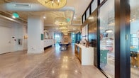 Located at 1017 Lake Ave in Lake Worth Beach, Florida, this single-floor coworking space blends tropical-inspired design with modern functionality. The welcoming reception area serves as the centerpiece, featuring a striking teal-tiled front desk adorned with a bold tropical mural backdrop and a glowing neon sign overhead. Woven rattan pendant chandeliers hang throughout the open interior, adding warmth and texture to the space's industrial-chic aesthetic, which includes exposed white ductwork and polished concrete floors.

The main corridor leads past a communal work area with a large shared table, geometric-patterned area rugs, and upholstered seating. A wall of numbered mailboxes provides practical postal services for members. Private glass-walled offices and enclosed meeting rooms offer focused work environments, benefiting from large steel-framed windows that invite abundant natural light and views of the outdoors.

A cozy lounge nook furnished with rattan chairs and a built-in bench provides an intimate setting for informal meetings or quiet work. Throughout the space, thoughtful design details — from decorative lighting to curated artwork — create an energetic yet comfortable atmosphere well-suited for professionals seeking a dynamic and inspiring place to work in South Florida.
