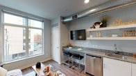 Located on Weston Street SE in the heart of downtown Grand Rapids, Michigan, this single-floor studio unit delivers a contemporary urban living experience with a thoughtfully designed open-concept layout. The space flows seamlessly from the living area to the sleeping quarters, all finished with warm wood-look flooring and cool gray walls that create a clean, modern aesthetic throughout.

An exposed metallic ductwork system runs along the ceiling, lending the space a distinctive industrial character. The kitchen area is efficiently outfitted with white cabinetry, granite-style countertops, stainless steel appliances including a dishwasher, refrigerator, and microwave, as well as an in-unit sink. Open floating shelves above the counter provide additional storage and display space, while a built-in breakfast bar with sleek bar seating and a wall-mounted flat-screen TV complete the area.

Large windows allow natural light to fill the interior and offer direct views of the surrounding downtown streetscape. The sleeping and living areas share an open, flexible space with recessed lighting overhead. A stacked washer and dryer unit is conveniently integrated into the layout. The bathroom features wood-look flooring, granite countertops, and a shower-tub combination, maintaining the consistent modern finishes found throughout the rest of the unit.