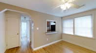 This single-floor residence offers a thoughtfully arranged layout designed for comfortable everyday living. The main living area is finished with warm hardwood flooring that extends throughout, creating a cohesive and inviting atmosphere. A ceiling fan with a light fixture provides both ventilation and ambient lighting, while a window fitted with blinds allows natural light to filter into the space.

A distinctive architectural detail is the pass-through window connecting the living area to the kitchen, framed in white with a granite ledge that serves as a convenient serving counter. The kitchen is visible through this opening and features rich, dark wood cabinetry paired with a black refrigerator, offering a classic and functional workspace.

A hallway leads from the main living area to the rest of the home, with smooth arched transitions between spaces that add a subtle design element. The hallway is well-lit with a ceiling fixture, and white trim and baseboards run throughout, complementing the neutral, warm-toned walls. The overall ambiance is clean, bright, and welcoming, with consistent finishes that give the space a unified and well-maintained character.