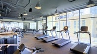 This contemporary fitness center is designed to offer an exceptional workout experience within a bright, open environment. The space is anchored by a row of commercial-grade treadmills positioned along expansive floor-to-ceiling windows, providing exercisers with abundant natural light and sweeping views of the surrounding cityscape. The industrial-chic aesthetic is underscored by exposed ductwork along the ceiling and bold pendant light fixtures that hang overhead, casting a focused yet inviting glow throughout the room. The flooring features durable rubber gym matting, purpose-built for high-traffic fitness use. Along the perimeter, a well-stocked free weight section includes a full rack of dumbbells and dedicated benching areas, catering to a range of strength training needs. Additional cardio and resistance equipment is thoughtfully arranged to maximize both functionality and flow through the space. Large mirrors line the walls, enhancing the sense of openness while allowing users to monitor their form. The overall ambiance is clean, modern, and energizing — a polished facility that strikes the ideal balance between professional-grade functionality and an inviting, light-filled atmosphere.
