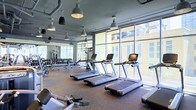 This contemporary fitness center is designed to offer an exceptional workout experience within a bright, open environment. The space is anchored by a row of commercial-grade treadmills positioned along expansive floor-to-ceiling windows, providing exercisers with abundant natural light and sweeping views of the surrounding cityscape. The industrial-chic aesthetic is underscored by exposed ductwork along the ceiling and bold pendant light fixtures that hang overhead, casting a focused yet inviting glow throughout the room. The flooring features durable rubber gym matting, purpose-built for high-traffic fitness use. Along the perimeter, a well-stocked free weight section includes a full rack of dumbbells and dedicated benching areas, catering to a range of strength training needs. Additional cardio and resistance equipment is thoughtfully arranged to maximize both functionality and flow through the space. Large mirrors line the walls, enhancing the sense of openness while allowing users to monitor their form. The overall ambiance is clean, modern, and energizing — a polished facility that strikes the ideal balance between professional-grade functionality and an inviting, light-filled atmosphere.